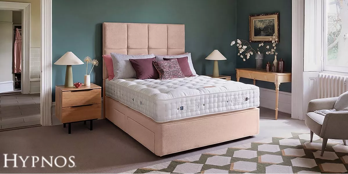 A plush tufted mattress on a light pink upholstered divan bed, styled with plum pillows in a bedroom with teal walls, wooden nightstands, matching table lamps, and a console table.